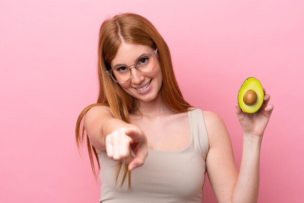 Mujer joven pelirroja sosteniendo un aguacate aislado sobre fondo rosa apuntando al frente con expresión feliz