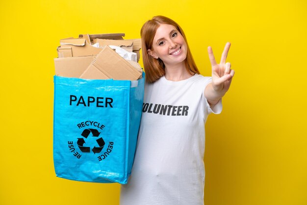 Mujer joven pelirroja sosteniendo una bolsa de reciclaje llena de papel para reciclar aislado sobre fondo amarillo sonriendo y mostrando el signo de la victoria