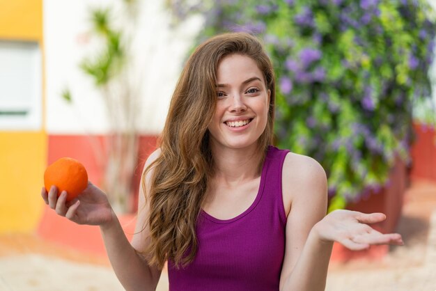 Mujer joven pelirroja sosteniendo una naranja al aire libre con expresión facial impactada