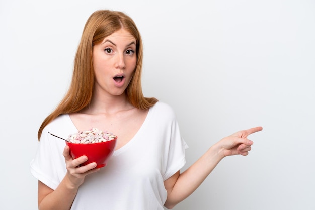 Mujer joven pelirroja sosteniendo un plato de cereales aislado sobre fondo blanco sorprendido y apuntando hacia el lado