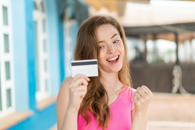 Mujer joven pelirroja sosteniendo una tarjeta de crédito al aire libre celebrando una victoria