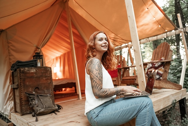 Mujer joven pelirroja con tatuaje usando laptop cerca de la tienda glamping. Vacaciones de estilo de vida de camping. Viajes de bajo presupuesto, viaje de vacaciones en el bosque. Tecnología de comunicación de información de conexión wifi.