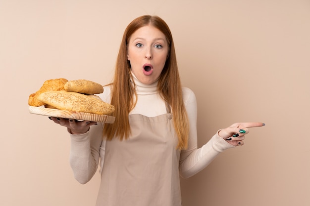 Mujer joven pelirroja en uniforme de chef. Panadero hembra sosteniendo una mesa con varios panes sorprendido y apuntando hacia el lado