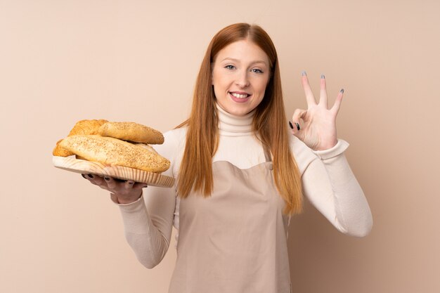 Mujer joven pelirroja en uniforme de chef. Panadero de sexo femenino que sostiene una tabla con varios panes que muestra la muestra aceptable con los dedos