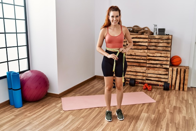 Mujer joven pelirroja usando cinta métrica que mide la cintura en el gimnasio luciendo positiva y feliz de pie y sonriendo con una sonrisa segura mostrando los dientes