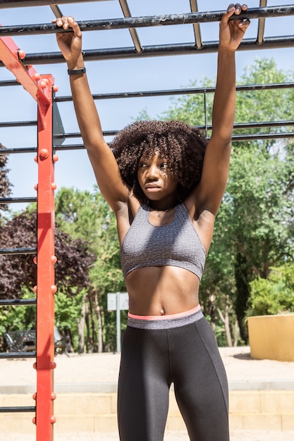 Mujer joven de pelo afro de pie en ropa deportiva negra y gris agarrándose a barras de calistenia negras y rojas con árboles verdes fuera de foco de fondo con iluminación natural dura