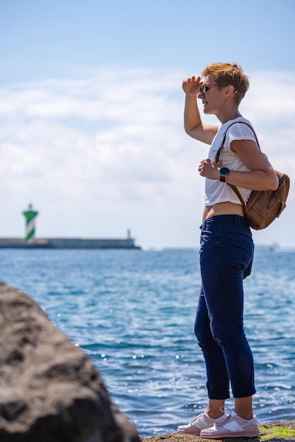 Mujer joven con pelo corto y mochila en la costa del mar Mediterráneo con faro y barcos en el fondo