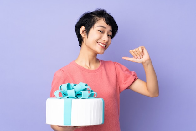 Mujer joven con pelo corto sosteniendo un gran pastel sobre púrpura orgulloso y satisfecho de sí mismo