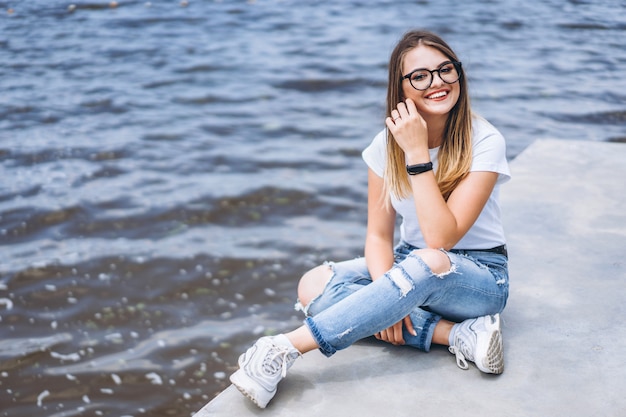 Mujer joven con el pelo largo en elegantes gafas posando en la orilla de hormigón cerca del lago. Niña vestida con jeans y camiseta sonriendo y mirando a la cámara