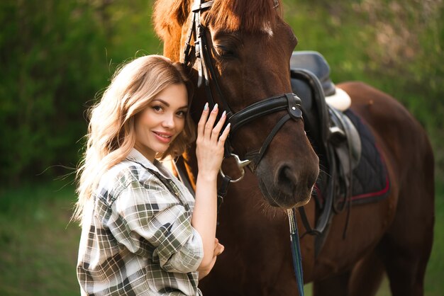 Mujer joven con pelo largo posando con un caballo marrón en un bosque en un prado soleado.