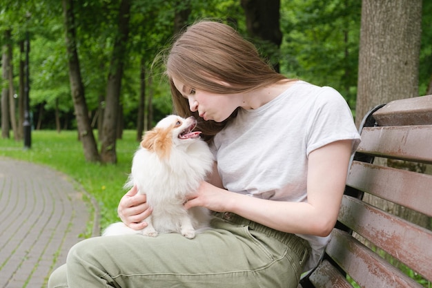 Mujer joven con el pelo largo y recto se sienta en un banco en el parque y besa a un perrito esponjoso en sus brazos