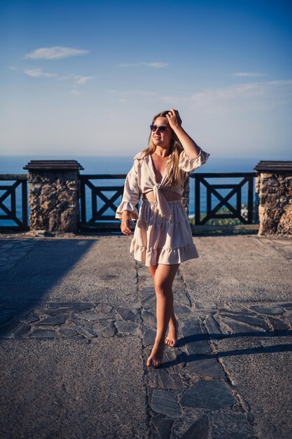 Una mujer joven con el pelo largo y rubio en un top y una falda camina en un día soleado de verano Una niña feliz con una sonrisa en la cara y gafas de sol camina en un día soleado