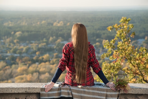 Mujer joven con el pelo largo se sienta en una colina con vistas al pueblo Vista posterior