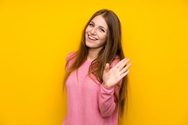 Mujer joven con el pelo largo sobre la pared amarilla aislada que saluda con la mano con la expresión feliz