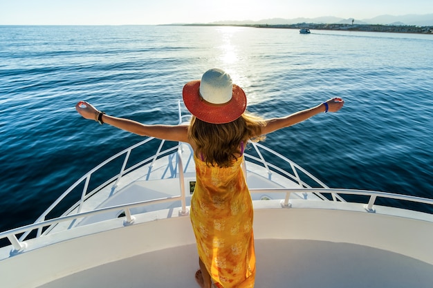 Mujer joven con el pelo largo con vestido amarillo y sombrero de paja de pie con las manos levantadas en la cubierta del yate blanco disfrutando de la vista del agua de mar azul