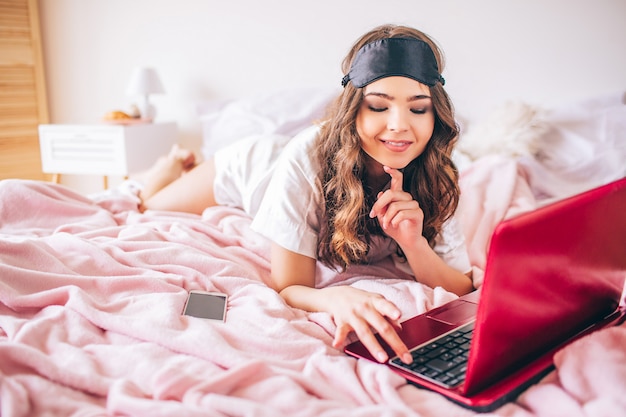 Mujer joven con el pelo oscuro acostado en cama en el dormitorio. Usando una computadora portátil y míralo. Modelo positivo alegre escribiendo en keybaord. Celular en la cama.