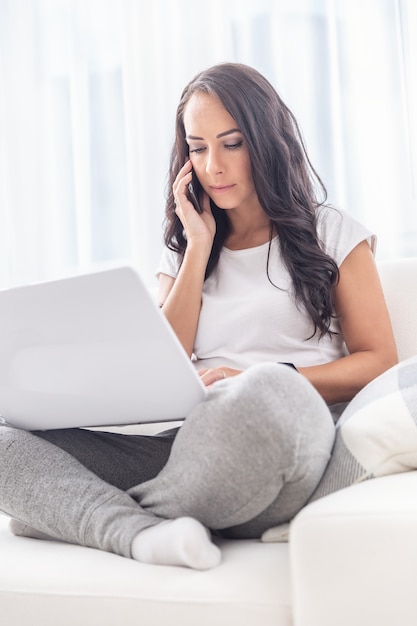 Mujer joven de pelo oscuro en la oficina en casa durante una cuarentena trabajando con su computadora portátil en su regazo, llamando con un cliente.
