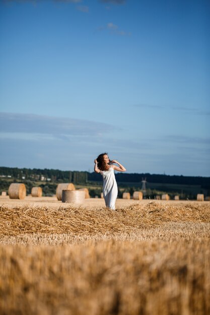 Mujer joven de pelo rizado en un campo de trigo, donde hay un enorme fajo de heno, disfrutando de la naturaleza. Personas y viajes. Naturaleza. rayos de sol agricultura