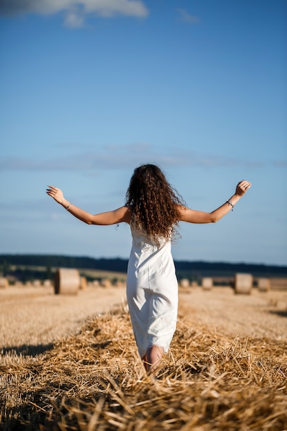 Mujer joven de pelo rizado en un campo de trigo, donde hay un enorme fajo de heno, disfrutando de la naturaleza. Personas y viajes. Naturaleza. rayos de sol agricultura