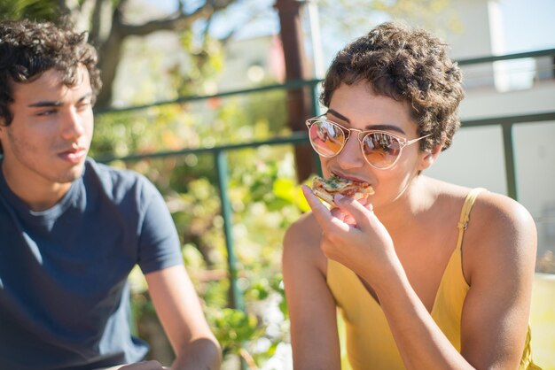 Mujer joven con pelo rizado en gafas de sol comiendo pizza sabrosa. Chica de pelo corto con vestido amarillo tomando un tentempié. Amigo en segundo plano. Fiesta, concepto de amigo