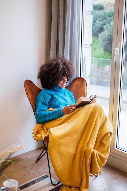 Mujer joven de pelo rizado leyendo un libro junto a la ventana cubierta por la manta disfrutando de su tiempo libre