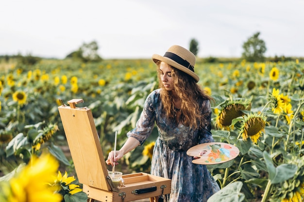 Una mujer joven con el pelo rizado y con un sombrero está pintando en la naturaleza. Una mujer se encuentra en un campo de girasol en un hermoso día