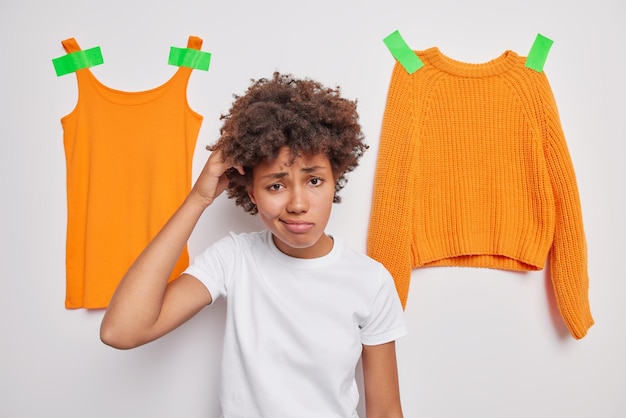 Mujer joven de pelo rizado vacilante desconcertada rasca la cabeza no sabe qué elegir vestida casualmente posa contra fondo blanco con camiseta y suéter enyesados. Concepto de personas y ropa.