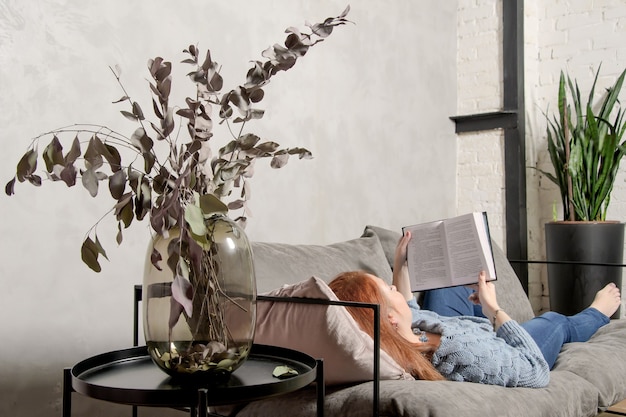 Foto mujer joven con pelo rojo está acostado en el sofá y leyendo un libro. casa de ocio tranquila con libro en interior acogedor.