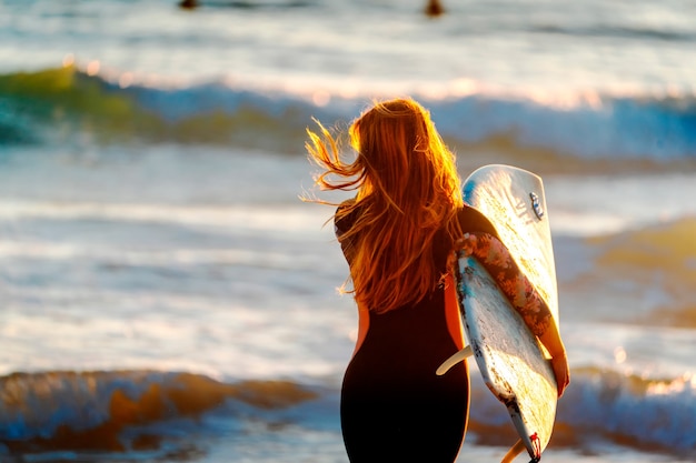 Mujer joven de pelo rojo caminando hacia el mar para surfear las olas al atardecer