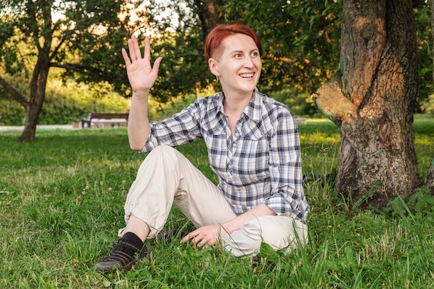 Mujer joven con pelo rojo corto se sienta en el césped del parque y saludando con la mano