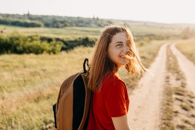 Una mujer joven con el pelo rojo y una mochila, caminando por un sendero, mirando a la cámara, sonriendo.