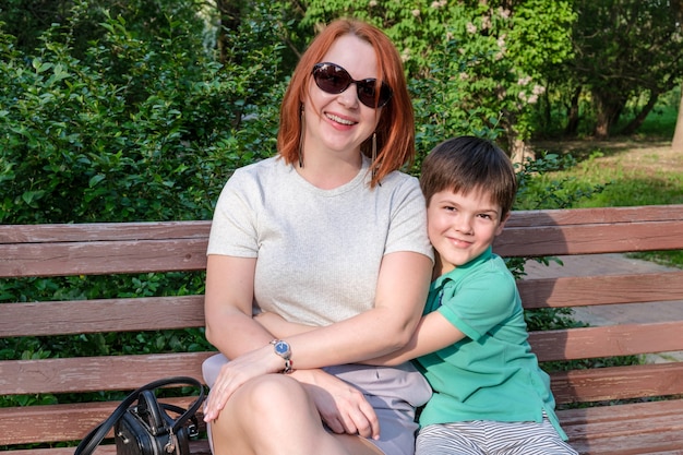 La mujer joven con el pelo rojo y el niño están sentados en el parque en un banco y sonríen. Hijo abraza a su madre suavemente. Concepto de crianza feliz, relación con los niños. Apego de los niños pequeños a su madre.