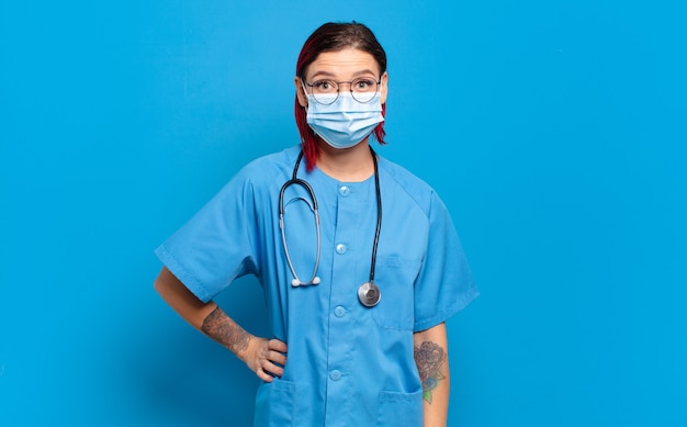 Mujer joven de pelo rojo sonriendo felizmente con una mano en la cadera y actitud confiada, positiva, orgullosa y amistosa. concepto de enfermera del hospital