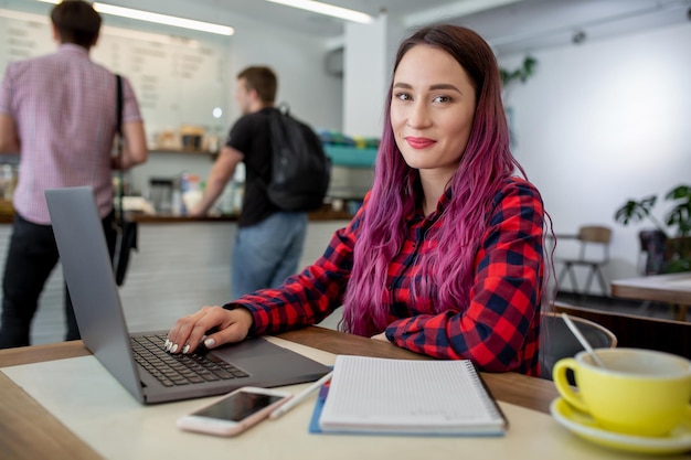 Mujer joven con el pelo rosa con ordenador portátil sentado en el café estudiante inteligente trabajando o ...