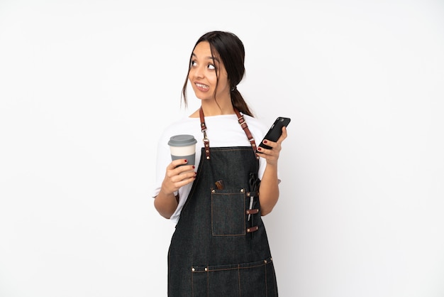 Mujer joven peluquera en blanco con café para llevar y un móvil mientras piensa algo