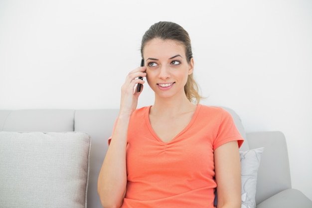 Mujer joven de pensamiento llamando con su teléfono inteligente sentado en el sofá