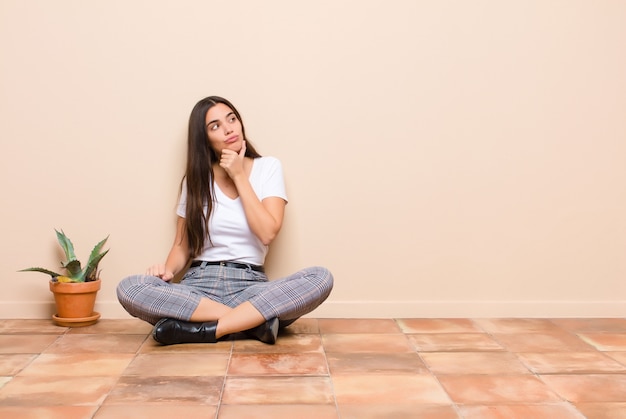 Mujer joven pensando, sintiéndose dudosa y confundida, con diferentes opciones, preguntándose qué decisión tomar sentado en el piso