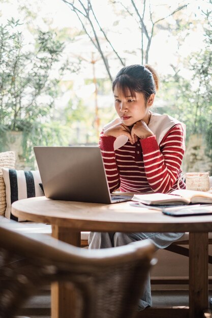 Mujer joven pensativa alejándose de su portátil en un momento contemplativo rodeada por el verde de un café brillante