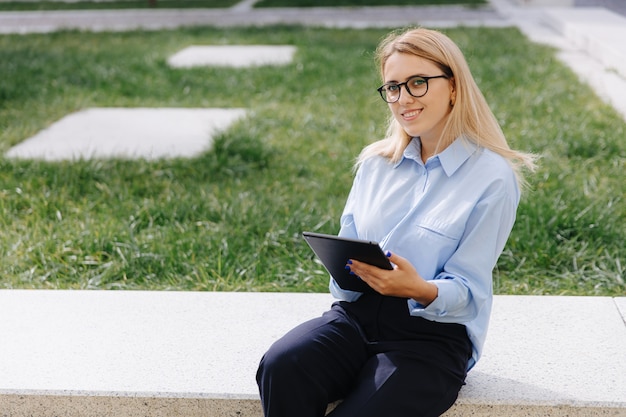 Mujer joven pensativa en camisa azul y jeans negros sentado al aire libre con tableta digital. Señora feliz con cabello rubio trabajando de forma remota en un dispositivo moderno.