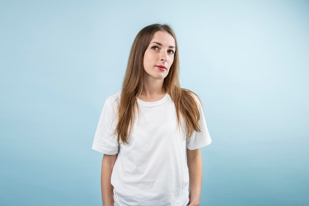 Mujer joven pensativa en camiseta blanca con pelo largo sobre fondo azul.