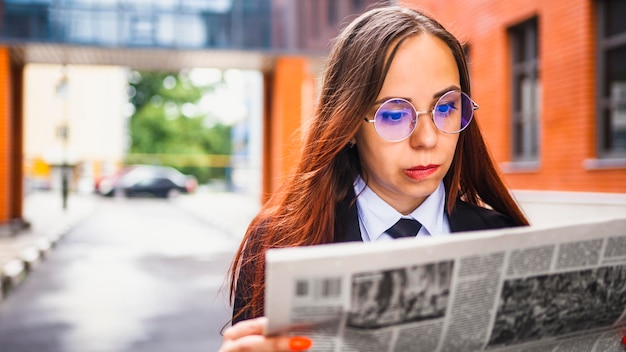 Mujer joven pensativa leyendo el periódico en la calle Empresaria joven confundida con cabello largo castaño en ropa formal y anteojos de pie en la calle de la ciudad leyendo el periódico