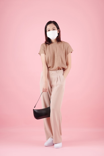 Mujer joven pensativa en máscara médica sosteniendo el bolso de cuero y poniendo la mano en el bolsillo