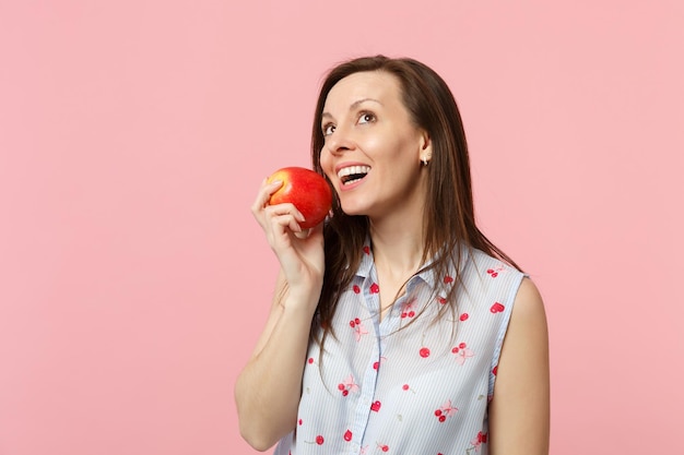 Mujer joven pensativa con ropa de verano mirando hacia arriba, sosteniendo fruta fresca de manzana roja madura aislada en un fondo rosa pastel en el estudio. El estilo de vida vívido de la gente relaja el concepto de vacaciones. Simulacros de espacio de copia.