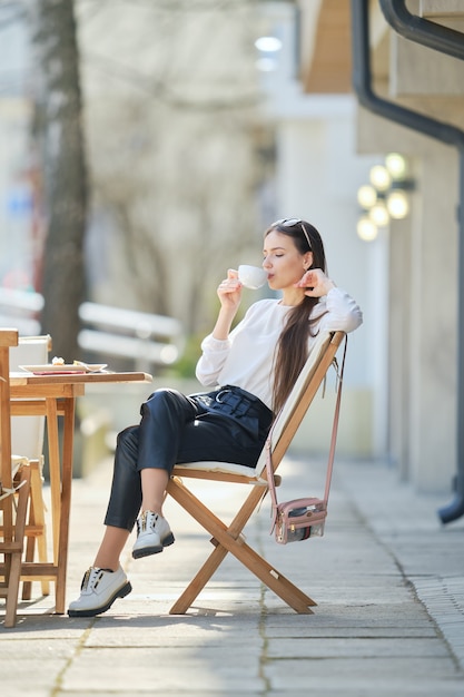 Mujer joven pensativa sentada en la terraza de la cafetería en un día soleado y tomando café.