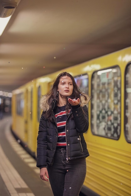 Mujer joven pensativa sosteniendo confeti mientras está de pie contra el tren en la estación de metro