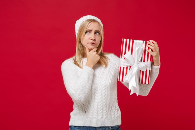 Mujer joven pensativa en suéter blanco, sombrero aislado sobre fondo de pared roja. Año Nuevo 2020, Día de San Valentín, cumpleaños, concepto de vacaciones. Simulacros de espacio de copia. Sostenga una caja de regalo a rayas con un lazo de cinta de regalo.