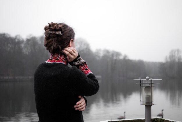 Foto una mujer joven está pensativa en la terraza en un día frío y mira hacia el lago gris