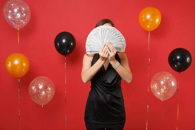 Mujer joven en pequeño vestido negro escondido, cubriendo la cara con un montón de dólares, dinero en efectivo en las manos sobre fondo rojo brillante globos de aire. Feliz año nuevo, concepto de fiesta de vacaciones de maqueta de cumpleaños.