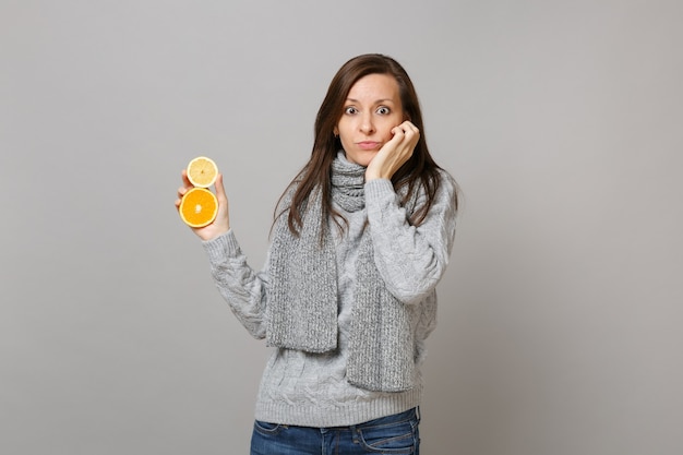 Mujer joven perpleja en suéter gris, bufanda puso la mano en la mejilla mantenga limón naranja aislado sobre fondo gris. Gente de estilo de vida de moda saludable emociones sinceras concepto de estación fría. Simulacros de espacio de copia.