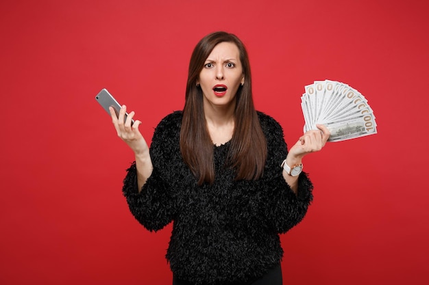 Mujer joven perpleja en suéter de piel negra con teléfono móvil, ventilador de dinero en billetes de dólar, dinero en efectivo aislado sobre fondo rojo. Concepto de estilo de vida de emociones sinceras de personas. Simulacros de espacio de copia.